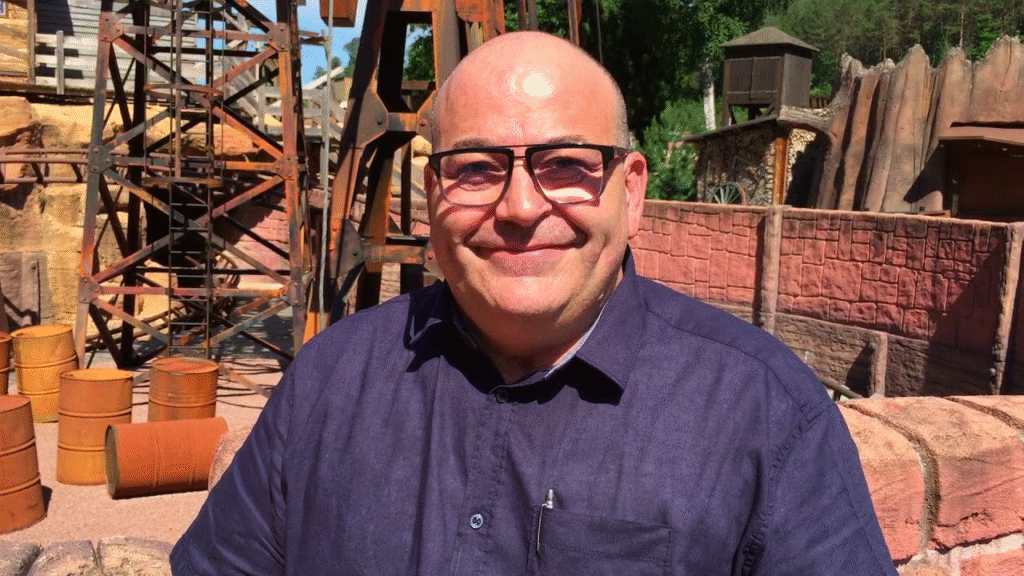 Portrait serré du directeur du parc : on distingue son visage souriant, coiffé de cheveux courts. Il regarde l’objectif, en extérieur, avec un fond évoquant l’univers western du parc.