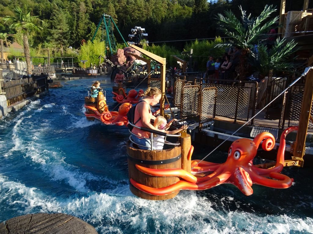 Sur la photo de L’île aux Pieuvres, on distingue une structure circulaire flottante en bois, ornée de tentacules géantes aux couleurs vives. Les passagers, installés dans des nacelles en forme de tonneaux, tiennent un volant pour contrôler leur proximité avec la rive.